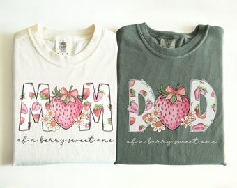 May include: Two t-shirts, one ivory and one moss green, with strawberry designs. The ivory shirt says "MOM" and the green shirt says "DAD", both with a strawberry graphic and the text "of a berry sweet one".
