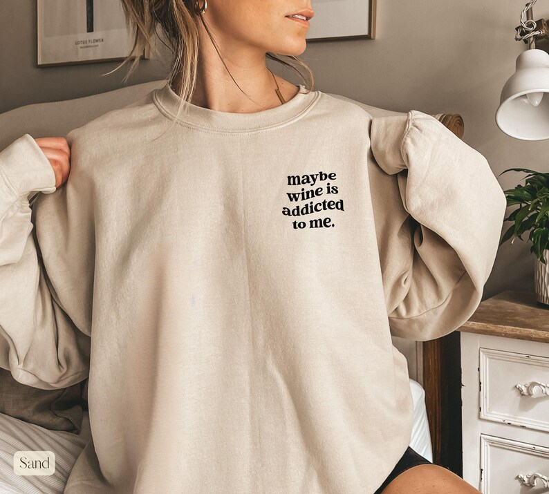 Puede incluir: Sudadera color arena con el texto "maybe wine is addicted to me." La parte superior de manga larga tiene un ajuste relajado y cuello redondo. El texto est&aacute; en una fuente negra simple.