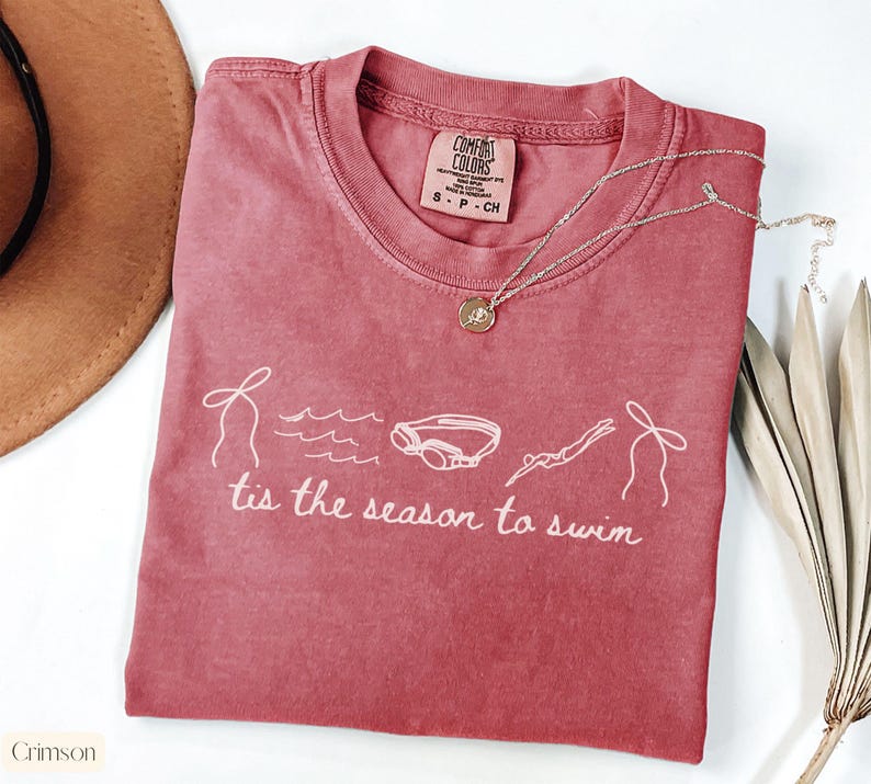 May include: Crimson-colored t-shirt with the phrase "tis the season to swim" and swimming-related graphics. The shirt has a crew neck and short sleeves. A gold necklace with a pendant is on the shirt.