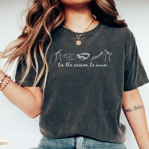May include: Dark gray t-shirt with white line art of a swimmer, goggles, and waves. The text "tis the season to swim" is printed below the graphic. The shirt is a relaxed fit.