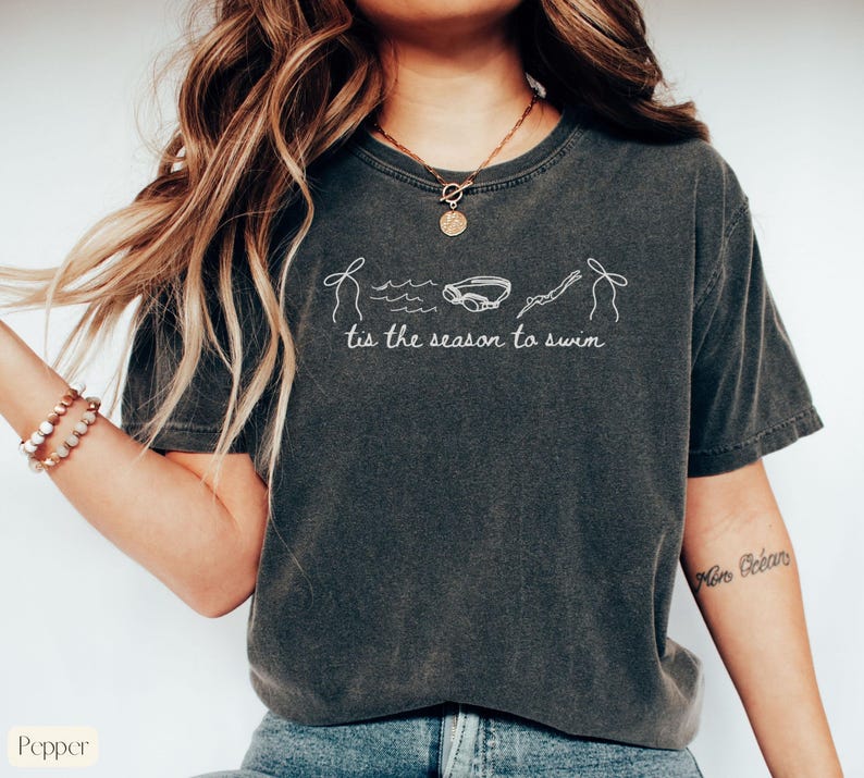 May include: Dark gray t-shirt with white line art of a swimmer, goggles, and waves. The text "tis the season to swim" is printed below the graphic. The shirt is a relaxed fit.