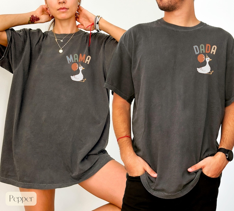 Puede incluir: Dos camisetas gris oscuro con dise&ntilde;os a juego. Una camiseta dice "MAMA" y la otra "DADA" encima de un ganso de dibujos animados que sostiene un globo. Las camisetas son de corte holgado y tienen mangas cortas.