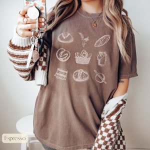 Peut inclure: Un t-shirt marron avec un motif blanc de diff&eacute;rents pains et articles de boulangerie. Le motif comprend un pain, un bretzel, un panier de pain, un pot de p&acirc;te, un rouleau &agrave; p&acirc;tisserie et une tige de bl&eacute;.