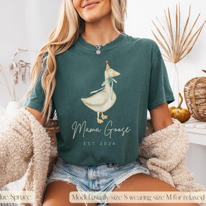 May include: A teal blue t-shirt with a white goose wearing a party hat and a blue ribbon. The text "Mama Goose EST 2024" is printed on the shirt.