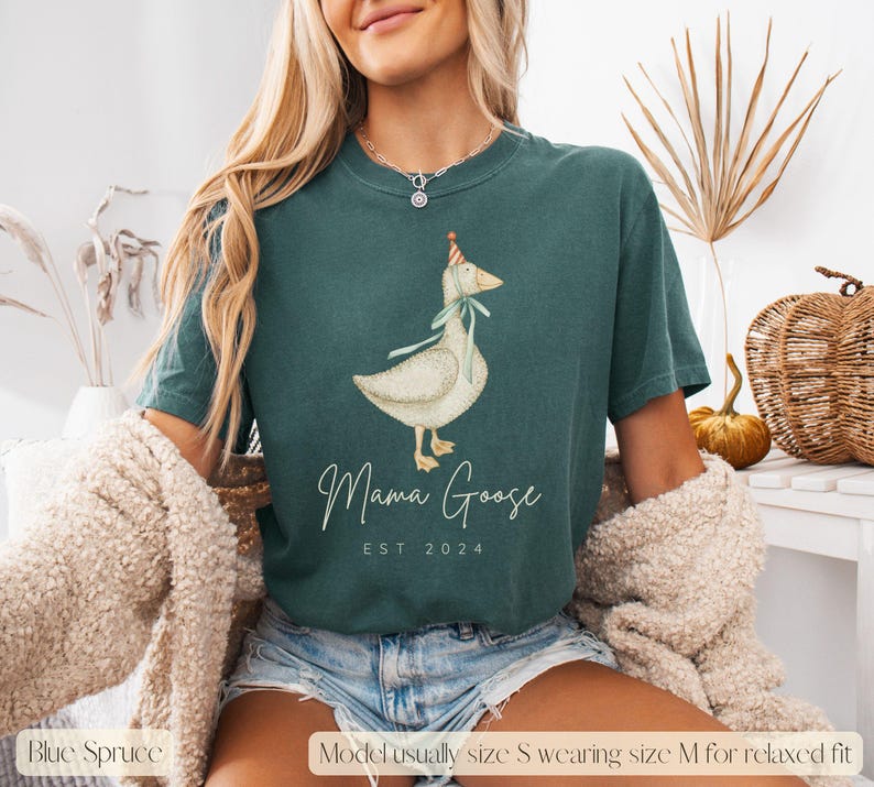 May include: A teal blue t-shirt with a white goose wearing a party hat and a blue ribbon. The text "Mama Goose EST 2024" is printed on the shirt.