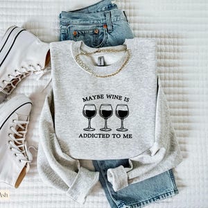 May include: A light gray sweatshirt with the text "Maybe Wine Is Addicted To Me" and three wine glasses. The sweatshirt is paired with blue jeans and white sneakers. A gold chain necklace is draped on the sweatshirt.