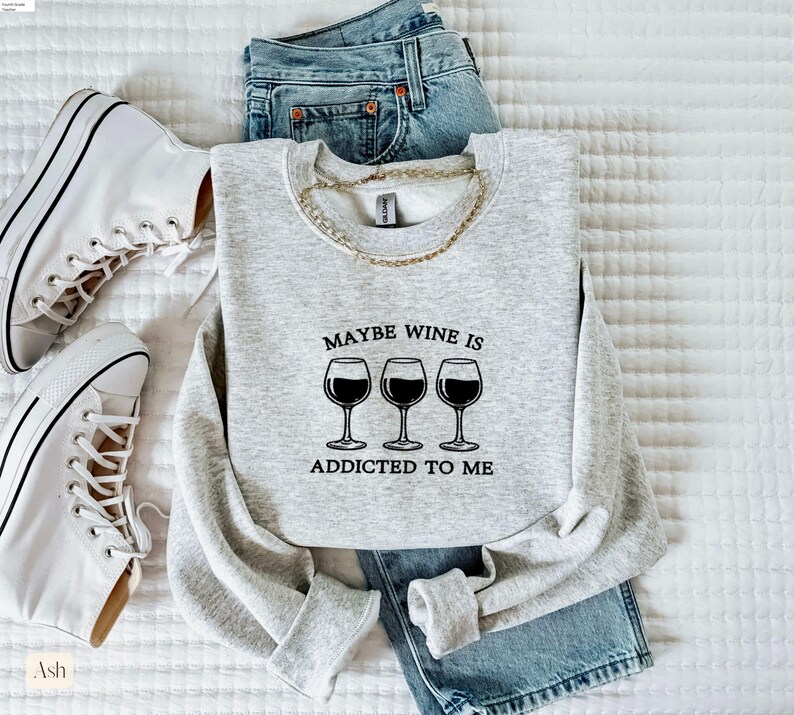 May include: A light gray sweatshirt with the text "Maybe Wine Is Addicted To Me" and three wine glasses. The sweatshirt is paired with blue jeans and white sneakers. A gold chain necklace is draped on the sweatshirt.