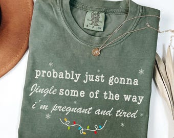Puede incluir: Una camiseta verde musgo con el texto blanco "probably just gonna Jingle some of the way i'm pregnant and tired". La camiseta tiene cuello redondo y mangas cortas. Un collar dorado con un colgante está sobre la camiseta.