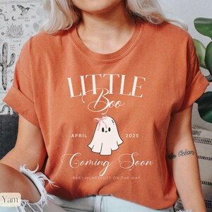 Op de afbeelding: Verbrand oranje t-shirt met de tekst "Little Boo Coming Soon" en een cartoon spook illustratie. De geest heeft een roze strik en de tekst "April 2025" staat onder de geest. De tekst "Baby Huneycutt on the way" staat onderaan.