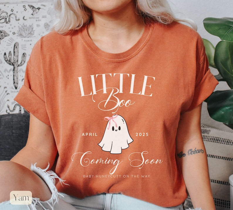 Op de afbeelding: Verbrand oranje t-shirt met de tekst "Little Boo Coming Soon" en een cartoon spook illustratie. De geest heeft een roze strik en de tekst "April 2025" staat onder de geest. De tekst "Baby Huneycutt on the way" staat onderaan.