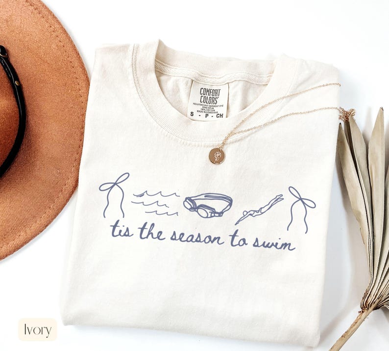 May include: Ivory-colored t-shirt with a blue graphic of swimming-related elements and the phrase "tis the season to swim." The shirt features a crew neck and short sleeves. A gold necklace with a pendant is draped on the shirt.