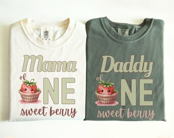 May include: Two t-shirts, one ivory and one moss green, with the text "Mama of One sweet berry" and "Daddy of One sweet berry" printed on them. Each shirt features a cartoon strawberry in a basket.