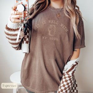 Puede incluir: Una camiseta marr&oacute;n de gran tama&ntilde;o con el texto "It is well with my soul." y un dibujo de un frasco con tapa.