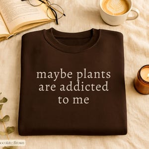 May include: Chocolate brown sweatshirt with the text "maybe plants are addicted to me" in white. The image also includes a lit candle, a cup of coffee, and an open book with glasses.