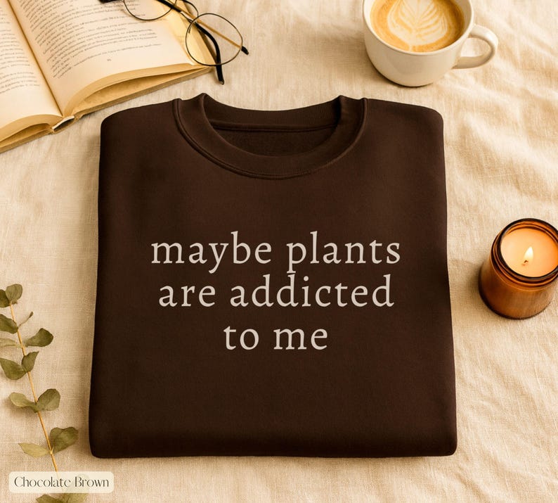 May include: Chocolate brown sweatshirt with the text "maybe plants are addicted to me" in white. The image also includes a lit candle, a cup of coffee, and an open book with glasses.