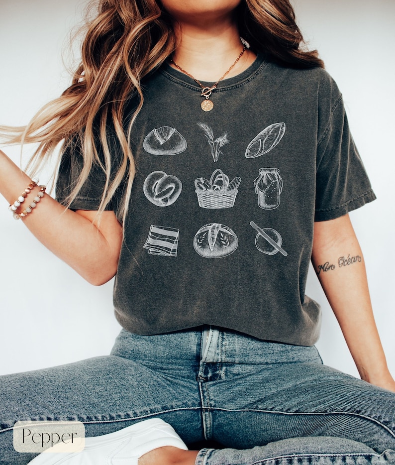 Peut inclure: Un t-shirt gris fonc&eacute; avec un imprim&eacute; graphique blanc de divers articles de boulangerie, notamment des pains, un rouleau &agrave; p&acirc;tisserie, un panier &agrave; pain et un pot de levain. Le texte "More Bread" est imprim&eacute; sur la manche droite du t-shirt.