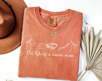 May include: A coral-colored t-shirt with the phrase "the life of a swim mom" in white script. The shirt features line drawings of swimming-related images. A gold necklace with a pendant is draped across the shirt.