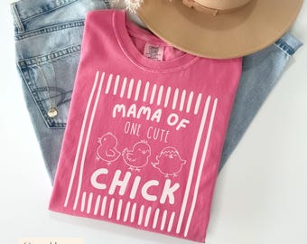 Op de afbeelding: Een roze t-shirt met de witte tekst "MAMA OF ONE CUTE CHICK" en drie cartoonkuikens. Het shirt is opgevouwen op een spijkerbroek, met een gouden ketting en een beige hoed.