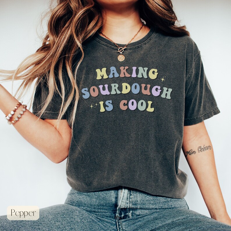 Peut inclure: Un t-shirt gris fonc&eacute; avec le texte "Making Sourdough is Cool" en couleurs arc-en-ciel. Le texte est dans un style de police r&eacute;tro.