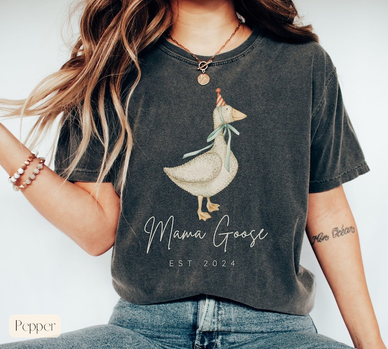 May include: A dark gray t-shirt with a white goose wearing a party hat and the text "Mama Goose EST 2024" printed on the front.