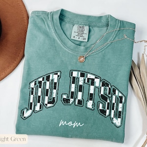 Pu&ograve; includere: T-shirt verde chiaro con la scritta "JIU JITSU" in un motivo a scacchi e la parola "mamma" in corsivo. La maglietta ha un girocollo e una piccola etichetta. Una collana d'argento con un ciondolo &egrave; drappeggiata sulla maglietta.