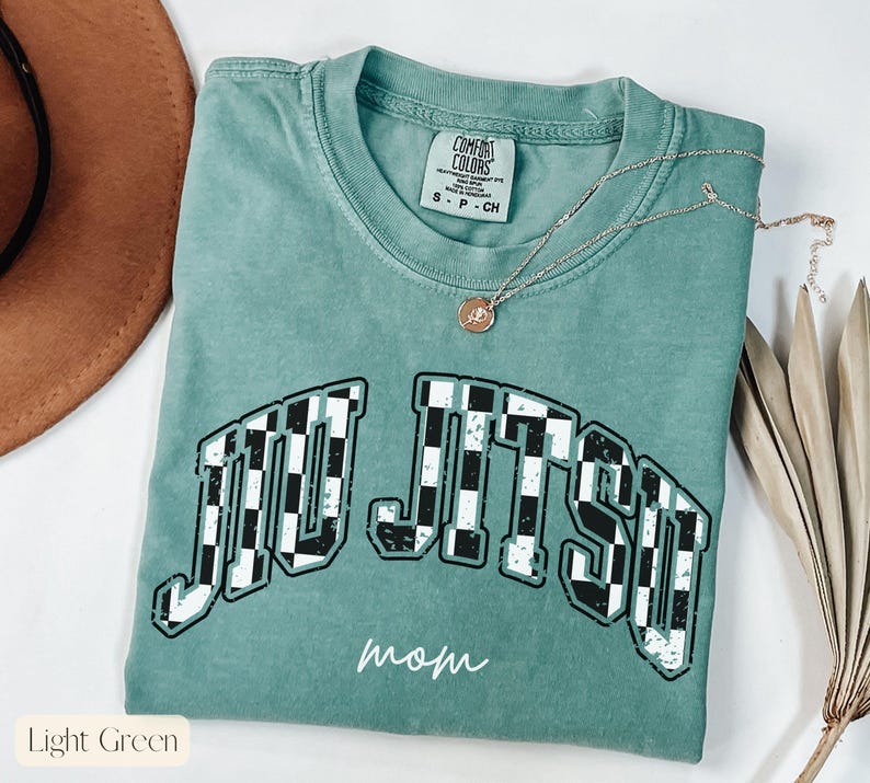 Pu&ograve; includere: T-shirt verde chiaro con la scritta "JIU JITSU" in un motivo a scacchi e la parola "mamma" in corsivo. La maglietta ha un girocollo e una piccola etichetta. Una collana d'argento con un ciondolo &egrave; drappeggiata sulla maglietta.