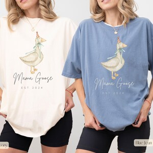May include: Two women wearing oversized t-shirts with a goose wearing a party hat and the text "Mama Goose Est. 2024". The woman on the left is wearing an ivory t-shirt and the woman on the right is wearing a blue jean t-shirt.