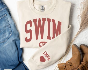May include: A sand-colored sweatshirt with the word "SWIM" in maroon block letters. The sleeve features the name "EMMA" and a heart. The sweatshirt is folded next to blue jeans and brown boots.