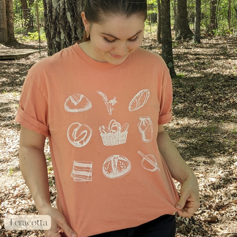 Peut inclure: Un t-shirt couleur terre cuite avec un motif graphique blanc de diff&eacute;rents pains et produits de boulangerie. Le motif comprend un pain, un bretzel, un panier de pain, une baguette, une p&acirc;tisserie et une poche &agrave; douille.
