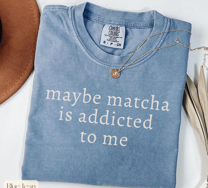 Puede incluir: Una camiseta azul vaquera Comfort Colors con el texto "maybe matcha is addicted to me" en blanco. Un collar dorado con un colgante est&aacute; drapeado sobre la camisa. La camisa es talla peque&ntilde;a.