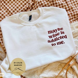 Puede incluir: Sudadera blanca con la frase bordada "maybe wine is addicted to me" en granate. La prenda se presenta sobre una superficie estampada, destacando la prenda informal.