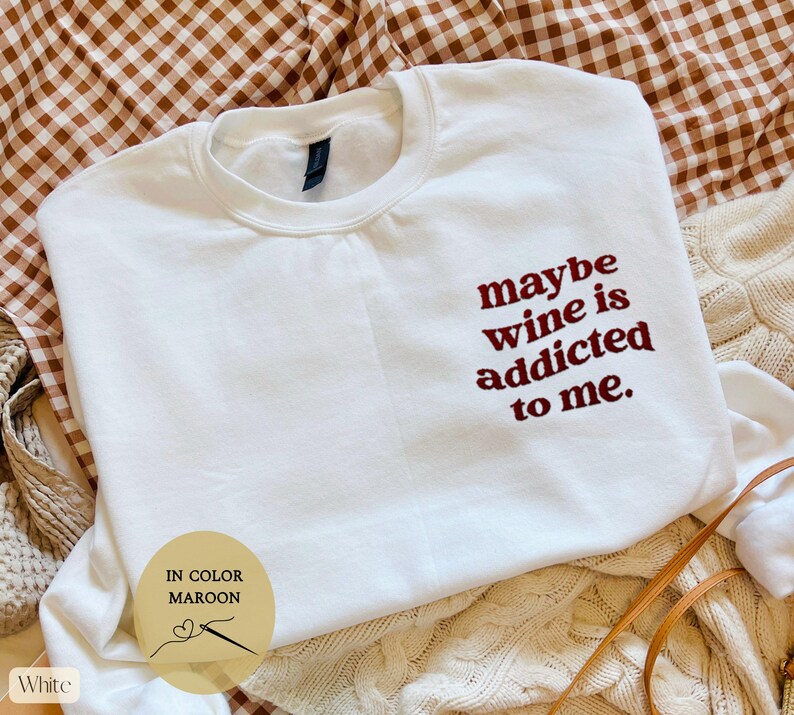 Puede incluir: Sudadera blanca con la frase bordada "maybe wine is addicted to me" en granate. La prenda se presenta sobre una superficie estampada, destacando la prenda informal.