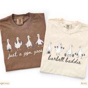 K&ouml;nnte beinhalten: Zwei T-Shirts, eines in Espresso und eines in Elfenbein, mit Rundhalsausschnitt. Das Espresso-Shirt hat den Text "just a gym goos" und das elfenbeinfarbene Shirt den Text "barbell baddie". Beide Shirts zeigen eine Grafik von G&auml;nsen, die Gewichte heben.