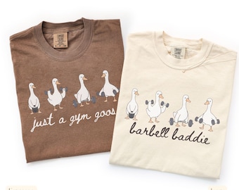 May include: Two t-shirts, one espresso and one ivory, with a crew neck. The espresso shirt has the text "just a gym goos" and the ivory shirt has the text "barbell baddie". Both shirts feature a graphic of geese lifting weights.