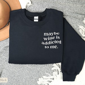 Puede incluir: Una sudadera negra con la frase bordada "maybe wine is addicted to me." La sudadera de cuello redondo est&aacute; doblada sobre una manta estampada, con un sombrero marr&oacute;n visible en el fondo. La sudadera es de la marca Gildan.