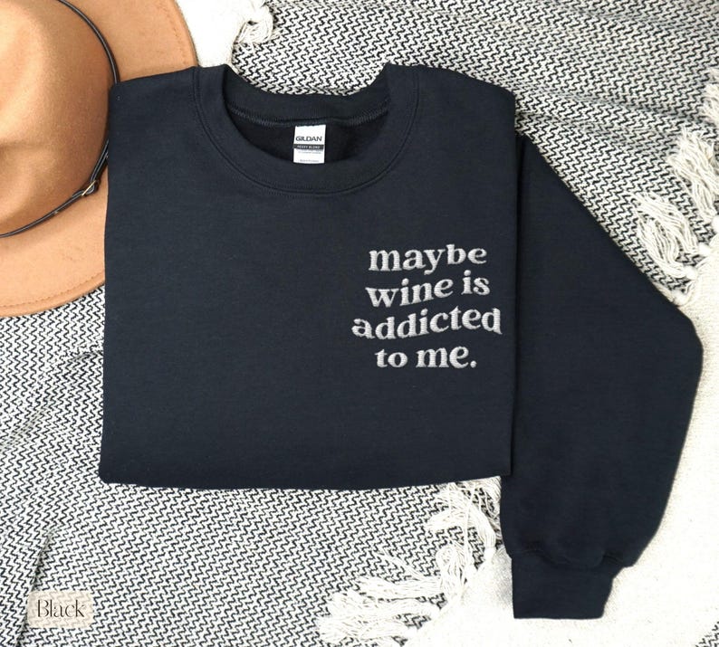 Puede incluir: Una sudadera negra con la frase bordada "maybe wine is addicted to me." La sudadera de cuello redondo est&aacute; doblada sobre una manta estampada, con un sombrero marr&oacute;n visible en el fondo. La sudadera es de la marca Gildan.