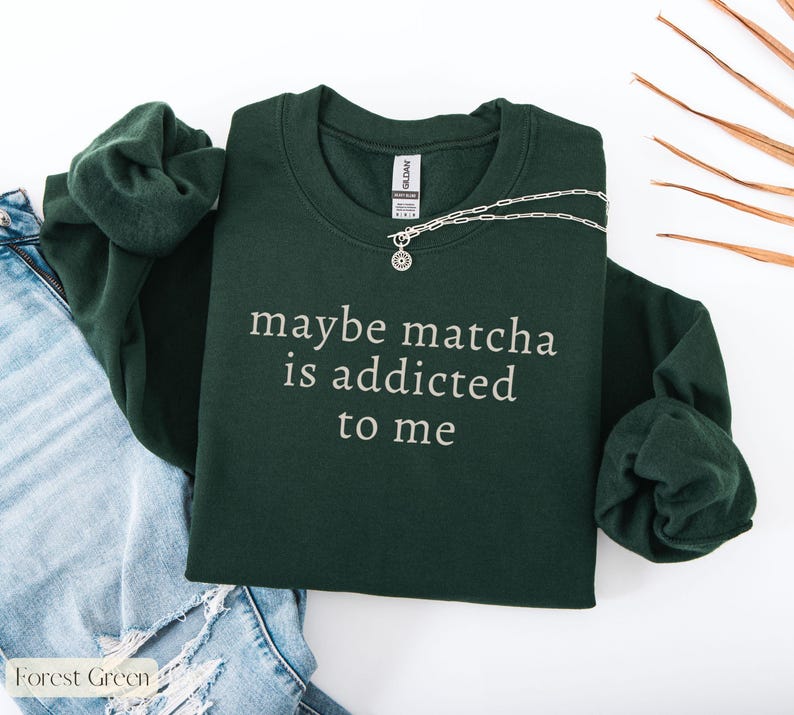Puede incluir: Sudadera verde bosque con el texto blanco "maybe matcha is addicted to me". Un collar plateado con un colgante est&aacute; colocado sobre la sudadera. La sudadera est&aacute; al lado de unos vaqueros azules.