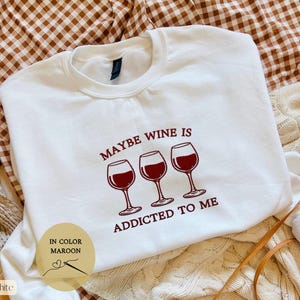 May include: White sweatshirt with the text "MAYBE WINE IS ADDICTED TO ME" in maroon. The design includes three wine glasses filled with red wine. The sweatshirt also has a small circular graphic that says "IN COLOR MAROON" and "White".