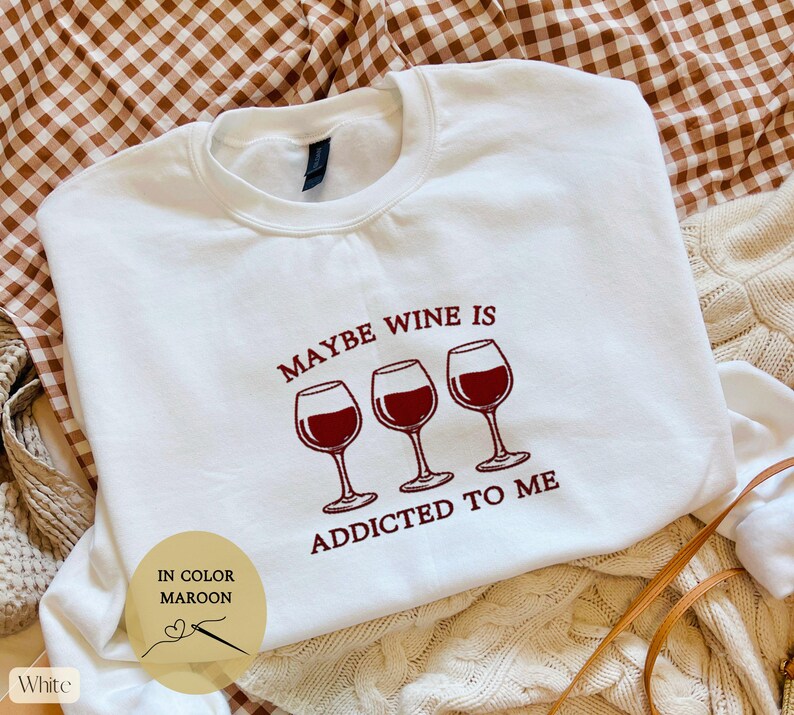 May include: White sweatshirt with the text "MAYBE WINE IS ADDICTED TO ME" in maroon. The design includes three wine glasses filled with red wine. The sweatshirt also has a small circular graphic that says "IN COLOR MAROON" and "White".