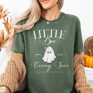 Op de afbeelding: Groen t-shirt met de tekst "LITTLE Boo" en een cartoon spookje met een roze strik. Op het shirt staat ook "April 2025" en "Coming Soon". De woorden "Baby Huneycutt on the way" staan ook op het shirt.