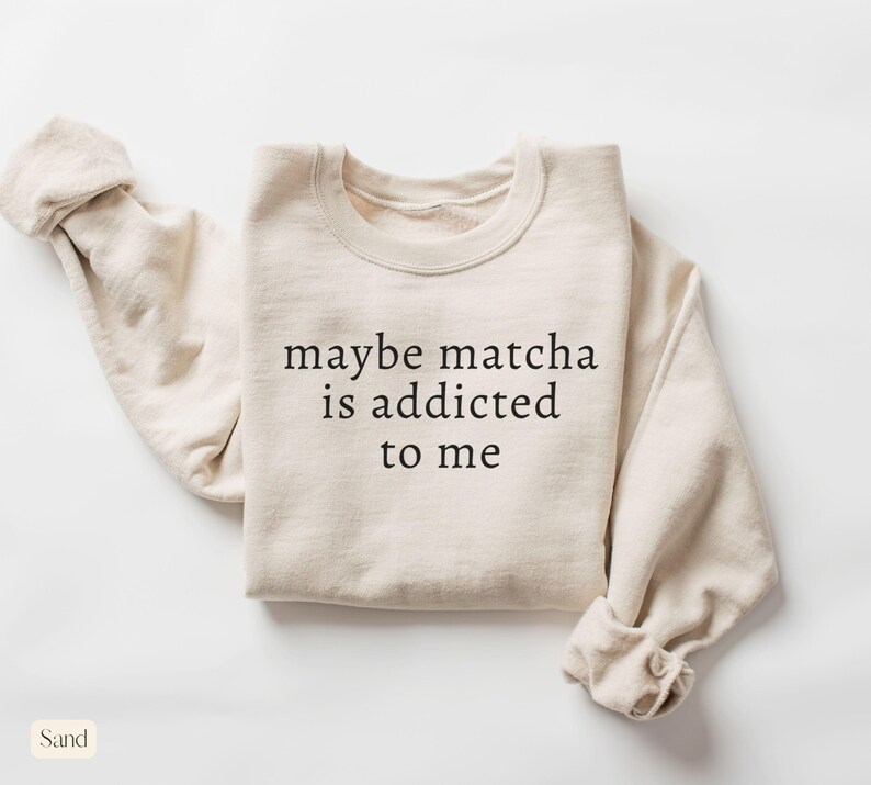 Puede incluir: Sudadera color arena con el texto negro "maybe matcha is addicted to me". La sudadera de cuello redondo tiene mangas largas, una de ellas enrollada. La palabra "Sand" est&aacute; en la esquina inferior izquierda.