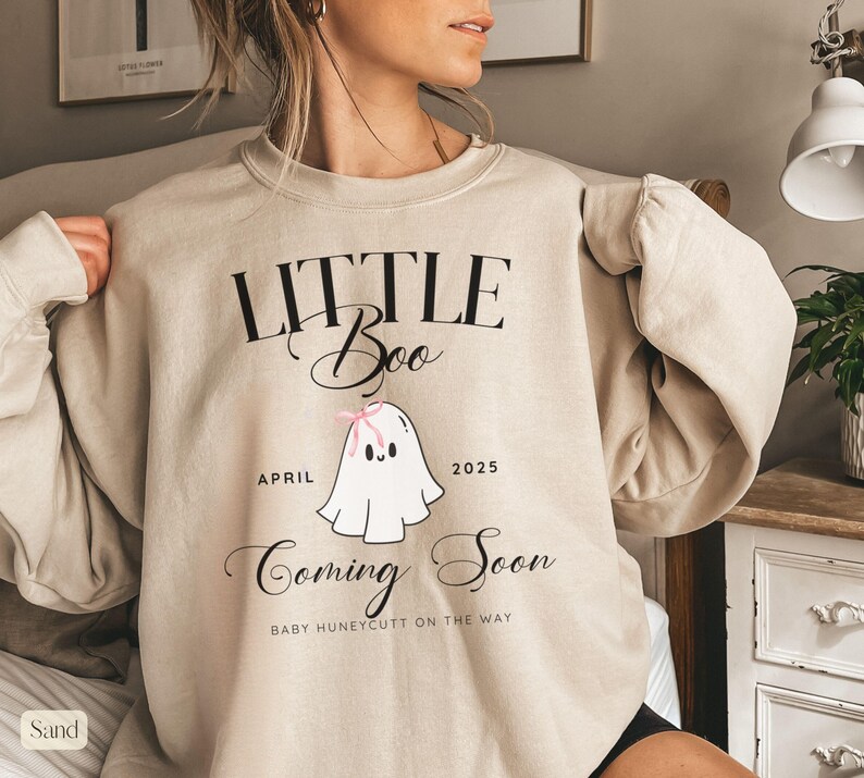 Op de afbeelding: Zandkleurige sweatshirt met de tekst "LITTLE Boo" boven een cartoongeest met een roze strik. Onder de geest staan de woorden "APRIL 2025" en "Coming Soon". De tekst "BABY HUNEYCUTT ON THE WAY" staat onder de woorden "Coming Soon".