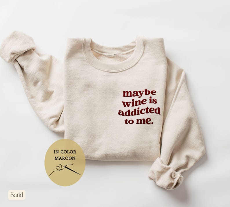 Puede incluir: Sudadera de color arena con la frase bordada "maybe wine is addicted to me" en granate. La sudadera tiene cuello redondo y mangas largas. Un gr&aacute;fico circular indica que el color es granate.