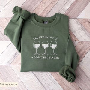 May include: Military green sweatshirt with the text "MAYBE WINE IS ADDICTED TO ME" and three wine glasses in white. The sweatshirt has a crew neck and long sleeves.