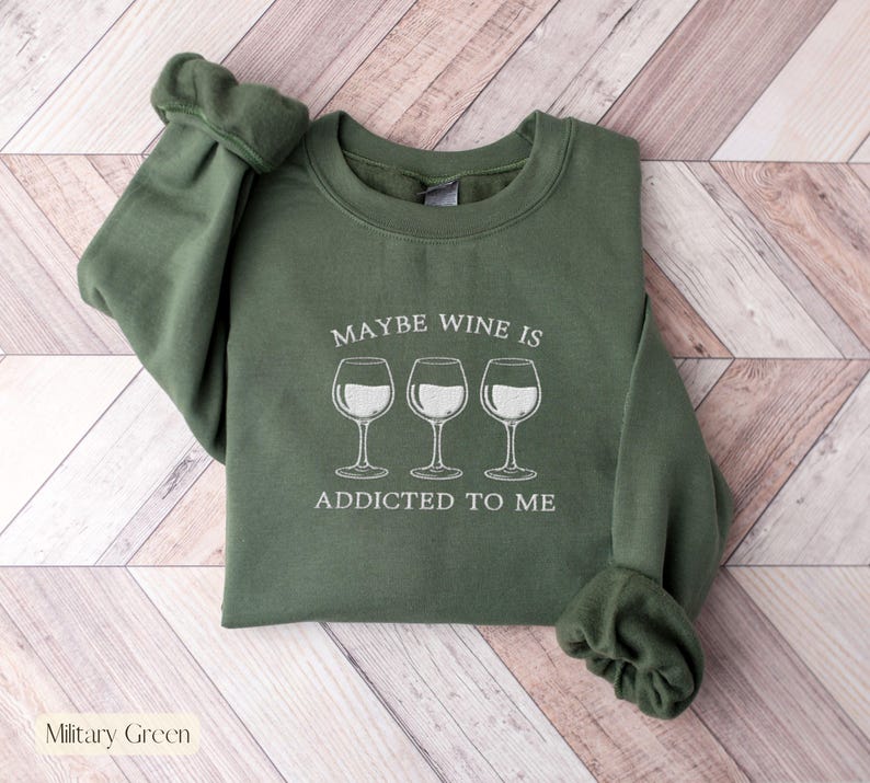 May include: Military green sweatshirt with the text "MAYBE WINE IS ADDICTED TO ME" and three wine glasses in white. The sweatshirt has a crew neck and long sleeves.