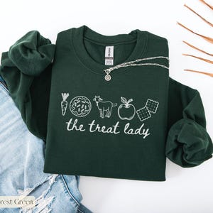 Puede incluir: Sudadera verde bosque con un gr&aacute;fico blanco de una zanahoria, una rosquilla, una cabra, una manzana y galletas saladas. El texto "the treat lady" est&aacute; debajo de los gr&aacute;ficos. Un collar plateado con un colgante est&aacute; sobre la sudadera. Se ven unos vaqueros azules en el fondo.