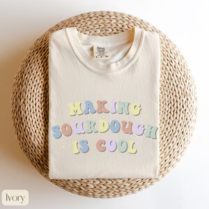 Peut inclure: Un t-shirt cr&egrave;me avec le texte "Making sourdough is cool" en lettres color&eacute;es de style r&eacute;tro. Le t-shirt est pli&eacute; et pr&eacute;sent&eacute; sur un tapis de paille tress&eacute;.