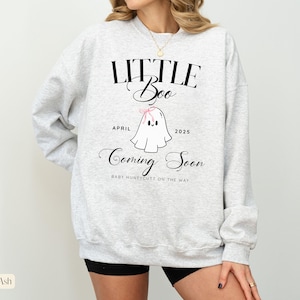 Op de afbeelding: Asgrijze sweatshirt met de tekst "LITTLE Boo" boven een cartoon spookje met een roze strik. De tekst "April 2025 Coming Soon" staat onder het spookje. De zin "BABY HUNEYCUTT ON THE WAY" staat onderaan.