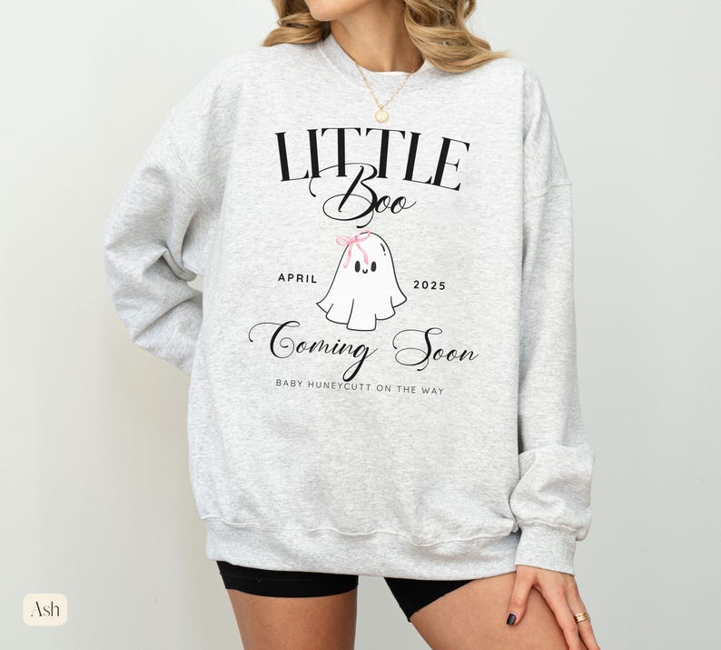 Op de afbeelding: Asgrijze sweatshirt met de tekst "LITTLE Boo" boven een cartoon spookje met een roze strik. De tekst "April 2025 Coming Soon" staat onder het spookje. De zin "BABY HUNEYCUTT ON THE WAY" staat onderaan.