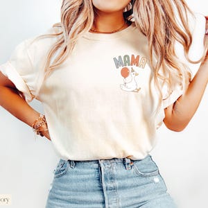 Puede incluir: Camiseta de color marfil con la palabra "MAMA" en letras pastel y un dibujo de un ganso sosteniendo un globo. El ganso lleva un gorro de fiesta. La camiseta se combina con vaqueros azules.